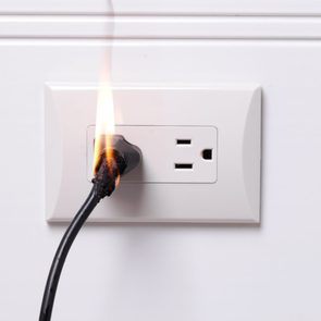 Electrical plug is burning in a wall outlet, causing flames and blackened wire in a plain white room.