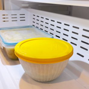 Round container with yellow lid sits inside refrigerator alongside rectangular container with blue lid, both on a white grated shelf.