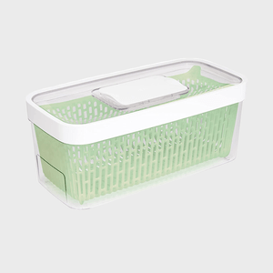 Transparent container holding a green perforated basket, resting on a white surface. The container has a white lid with a rectangular handle.