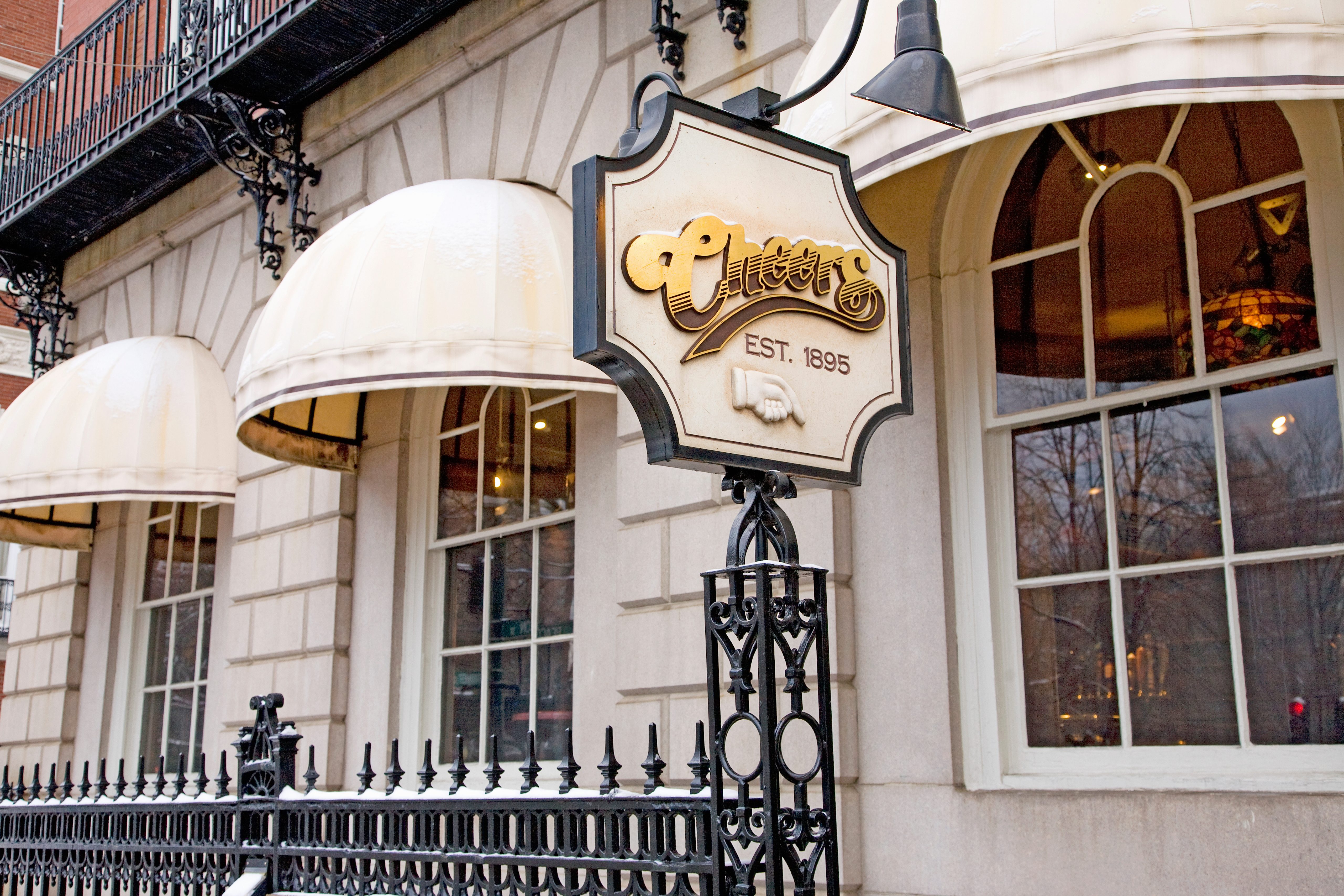 Sign reading "Cheers EST. 1895" is mounted on ornate ironwork in front of a building with arched windows and cream-colored awnings.