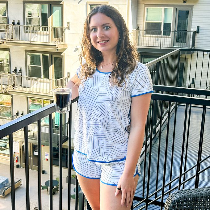 A person, holding a glass cup, stands on a balcony overlooking an apartment complex with visible railings and windows.