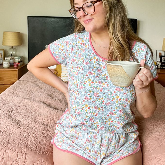 A person holds a mug, wearing floral pajamas, sitting on a textured bed. The room has bedside tables with lamps and decor.