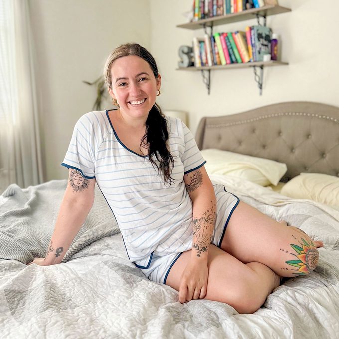 Person sitting on a bed, smiling, with tattoos visible on arms and legs; books and decor on shelves in the background.
