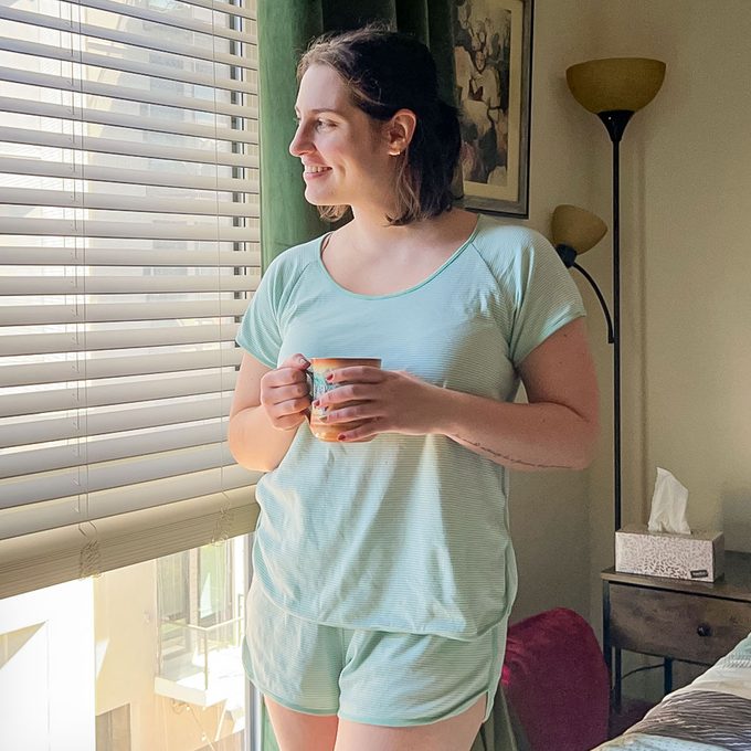 A person holds a mug, gazing out a window with blinds, in a cozy room featuring a lamp and a framed painting.
