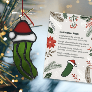 Pickle ornament hangs on tree; card beside describes a holiday tradition. Background features festive decorations with lights and pine branches.