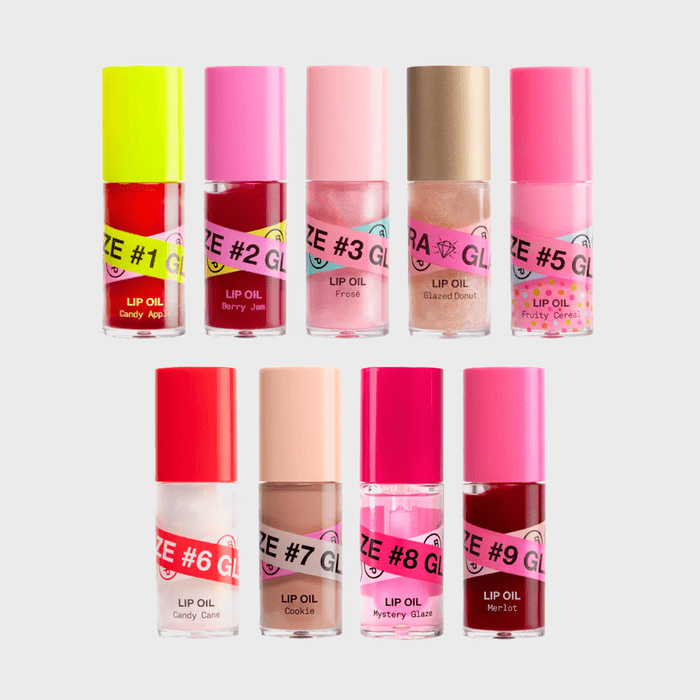 Various lip oil bottles stand arranged in rows, featuring colorful caps and labels with names like Candy Apple, Berry Jam, Cookie, and Merlot.