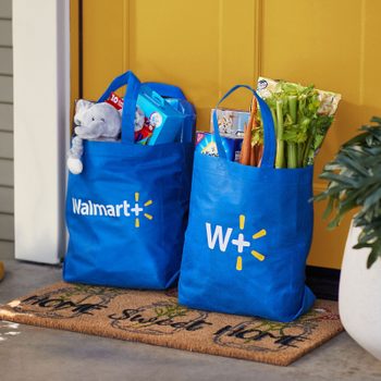 Blue Walmart+ bags sit filled with groceries and toys beside a yellow door, on a welcome mat near a potted plant.