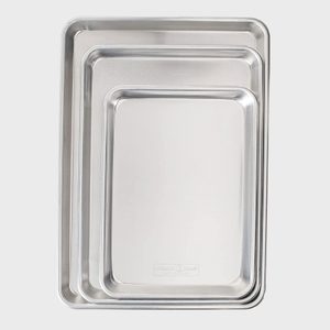 Three nested metal baking trays stacked in size order, sitting on a plain, light background. The smallest tray has a stamped logo.