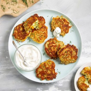 Zucchini fritters lie on a plate with a dollop of sour cream, garnished with dill, next to a wooden board and dish of sour cream.