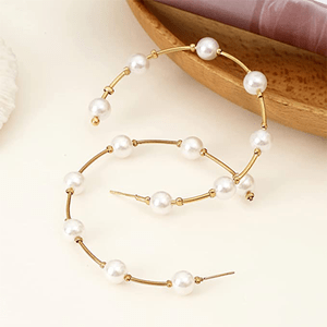 Hoop earrings with pearls connected by gold segments, resting on a light surface near a wooden bowl and cloth.
