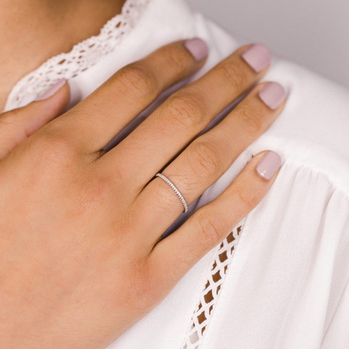 A hand rests on white lace fabric, wearing a slim, delicate ring; nails are painted in a pale, natural shade.