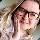 A person wearing glasses smiles gently, holding their cheek with a hand, in a well-lit room with neutral tones.