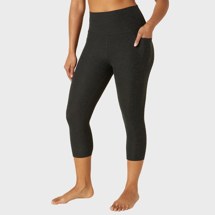 Person wearing dark leggings stands relaxed, posing with one hand in a pocket against a plain background.