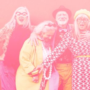 Group of smiling adults pose together, embracing joyfully against a vibrant pink background. Fashionable, colorful clothing and accessories enhance the lively atmosphere.
