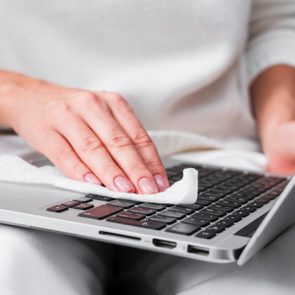 Hand cleans laptop keyboard with white cloth, seated in a cozy home environment.