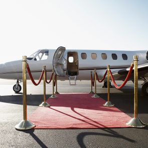 A private jet stands stationary on the tarmac, its entrance door open, framed by a red carpet and gold stanchions.