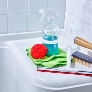 Cleaning supplies rest on a bathtub edge: spray bottle, red sponge, green cloth, squeegee, and scrub brush, with white tiled walls.