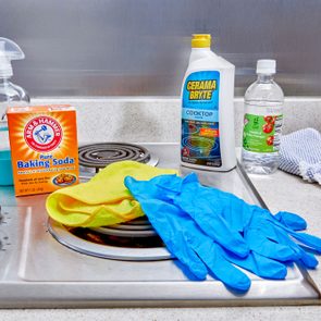 Cleaning supplies arranged on stove, including baking soda, spray bottle, gloves, and vinegar, with cloth next to stove knobs in a kitchen setting.