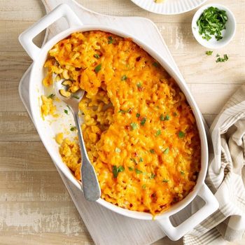 Baked macaroni and cheese is partially scooped with a spoon in a white dish, garnished with parsley, on a wooden table with a napkin nearby.