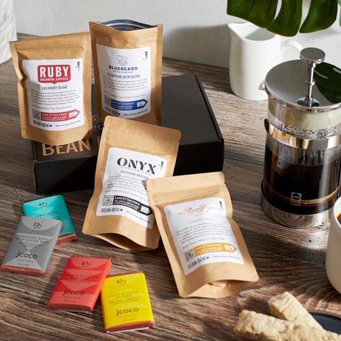 Coffee bags and chocolate bars rest beside a French press and a black box on a wooden table with a white pitcher nearby.