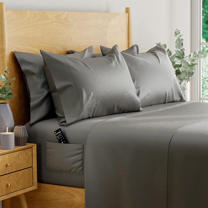 Gray bedding with a remote in pocket lies on a wooden bed, beside a nightstand with candles and eucalyptus in a vase.
