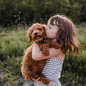 A child kisses a fluffy dog while holding it, surrounded by greenery in a field.