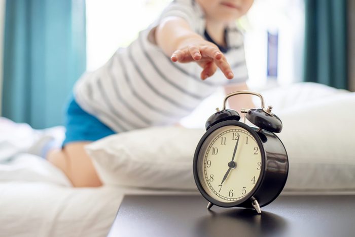 Sleepy little child lies on the bed and reaches for the ringing alarm clock. Sleepy boy on the bed with alarm clock in front.
