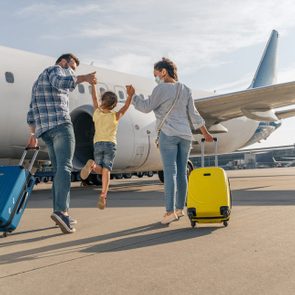 Family walking, holding hands, pulling suitcases on an airport tarmac towards a large airplane under a bright sky.