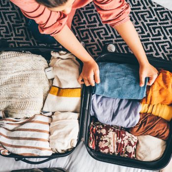 Open suitcase neatly packed with colorful clothes; person adjusting garments on a bed with a patterned blanket.