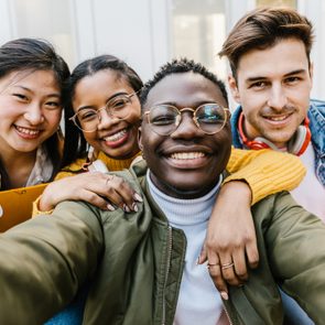 A group of four friends smiling, taking a selfie outdoors, with one person making a peace sign beside modern buildings.