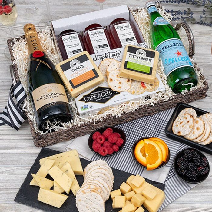 A wicker basket holds champagne, cheese, juice, and sparkling water, surrounded by raspberries, blackberries, orange slices, and crackers on a wooden surface.
