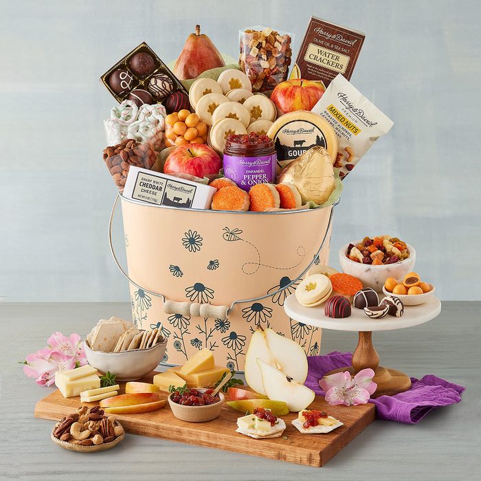 A decorative bucket holds assorted fruits, chocolates, and snacks on a wooden table surrounded by small dishes, flowers, and a purple cloth.