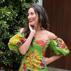Woman smiling, wearing a colorful floral dress, hand on cheek, standing in front of lush green plants and a wooden fence.
