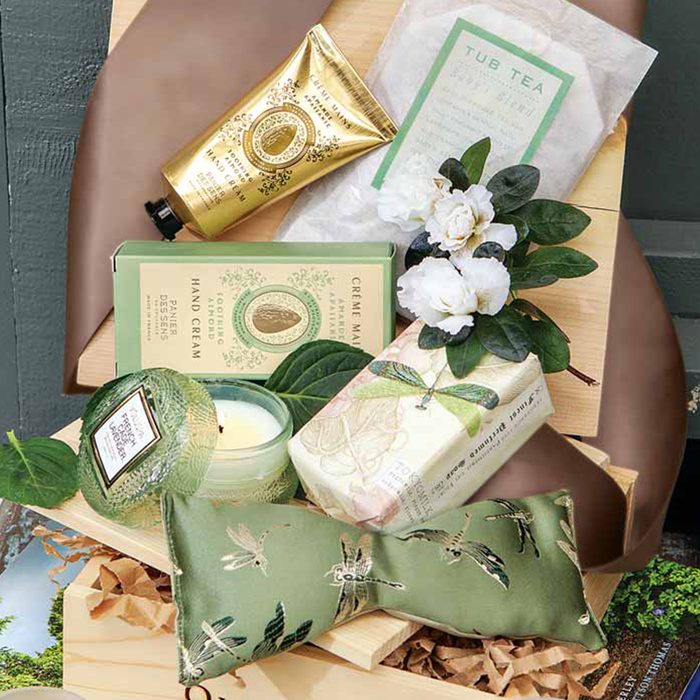 Luxurious toiletry items arranged in a wooden box; includes hand cream, soap, candle, tub tea, decorated with green leaves and white flowers.