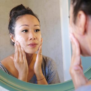 Woman applying skincare cream to her face, reflected in a round mirror, inside a bathroom setting.