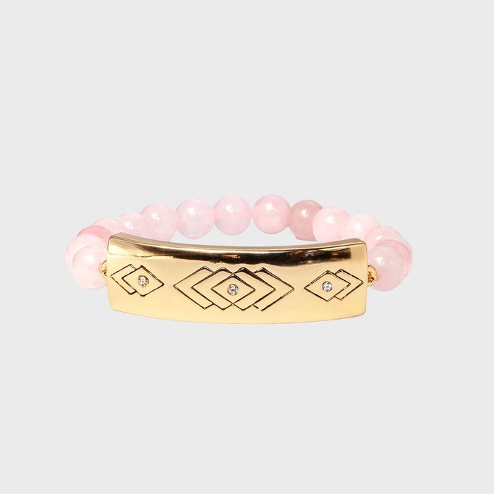 Gold bracelet with geometric engravings sits among pink beads against a plain gray background.