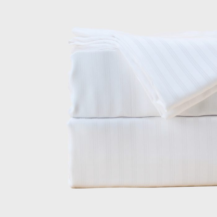 Folded white bedsheet resting on a flat surface, displaying faint vertical stripes; surrounded by a plain, pale background.