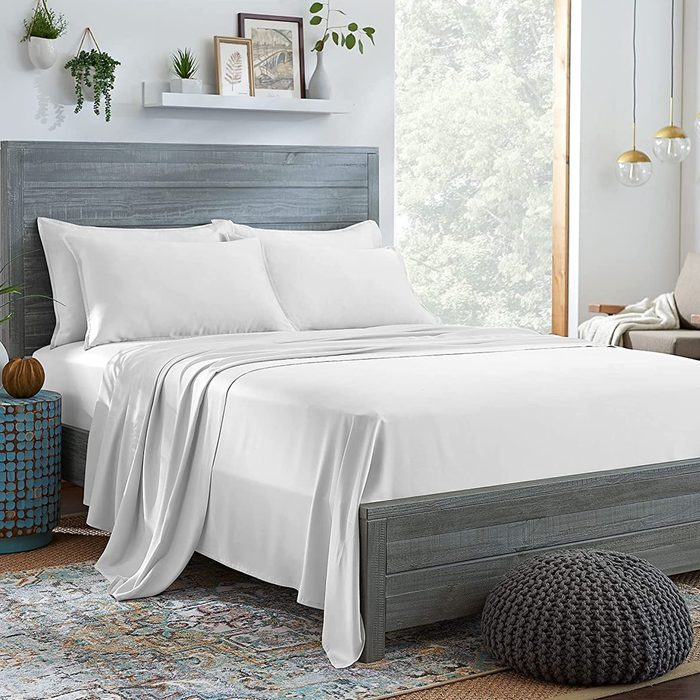 A neatly made bed with white sheets sits in a cozy bedroom with wall art, plants, and natural light from a large window.