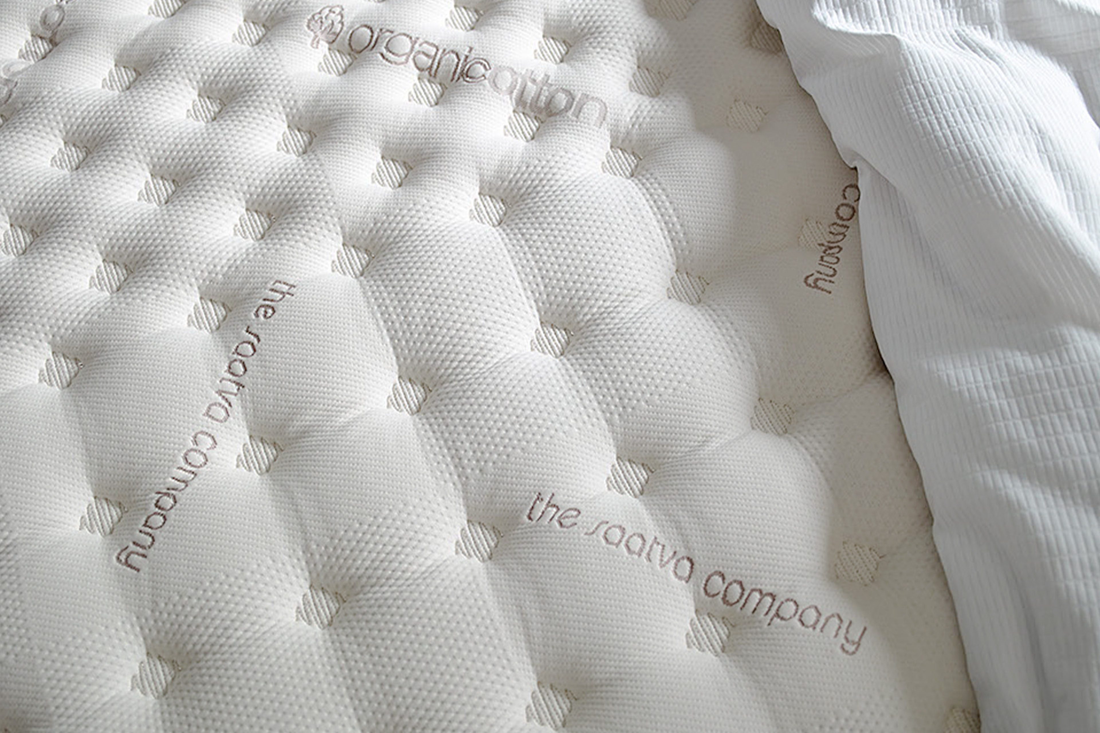 The mattress displays embroidered text "organic cotton, the saatva company" with a quilted pattern under soft lighting.