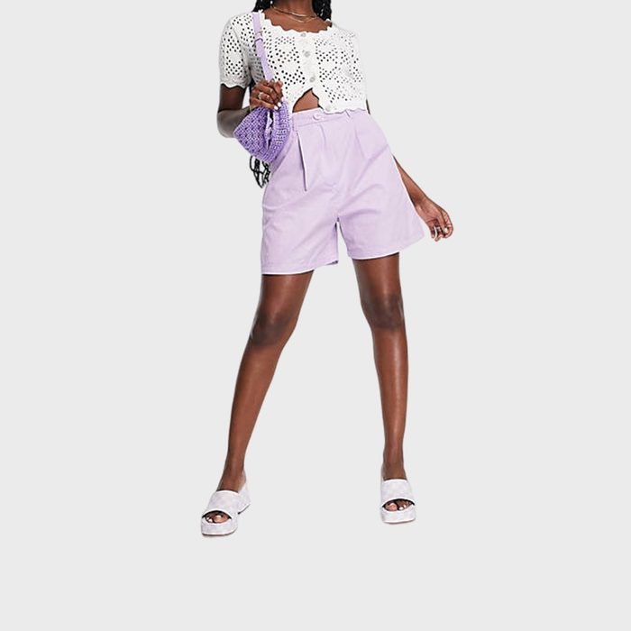 Person standing confidently, wearing a white crocheted top and lilac shorts, holding a purple bag. White sandals complete the casual outfit.