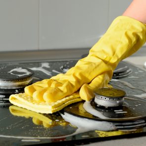 Hand in yellow glove cleaning stovetop with sponge, surrounded by soapy bubbles in a kitchen setting.