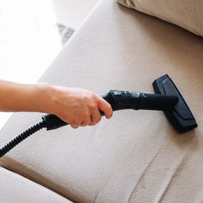 Vacuum nozzle cleans beige couch; hand grips handle; modern, tidy room.