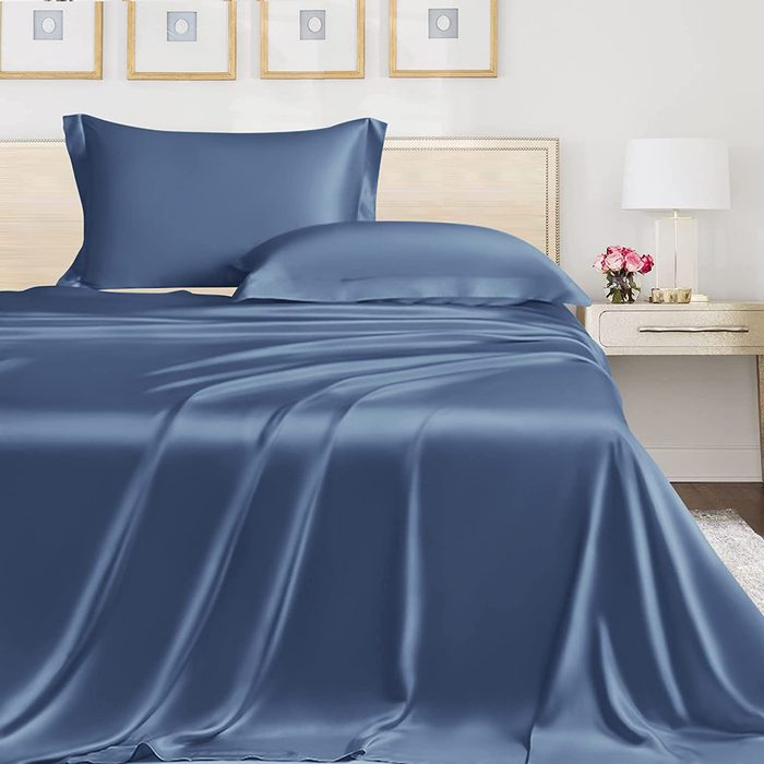 Blue satin bedding lies smoothly on a bed with wooden headboard, beside a nightstand with a lamp and flowers, in a minimalist bedroom.