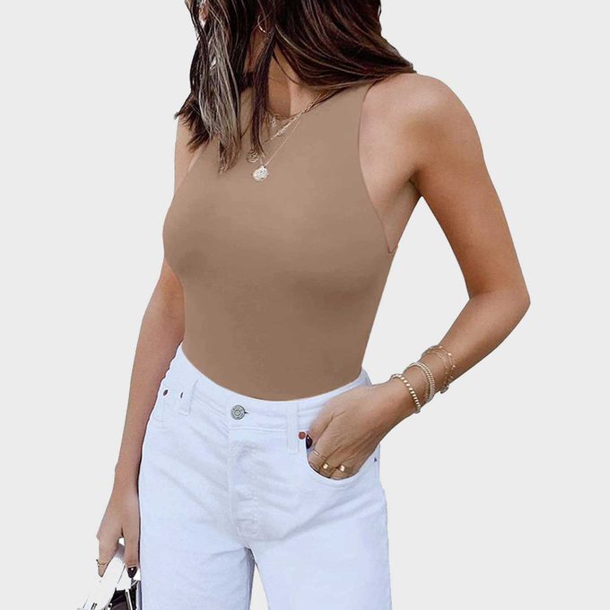 Person wears a beige tank top and white jeans, holding a purse. The individual stands against a plain white background, adorned with jewelry.