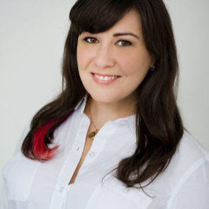 A person smiles softly, wearing a white shirt, with a pink streak in their dark hair, against a plain white background.