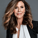 A person with long wavy hair smiles slightly, wearing a black blazer and white shirt, against a plain gray background.
