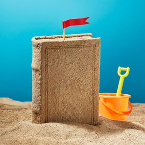A sand book stands vertically with a red flag; beside it, an orange bucket and yellow shovel sit on sandy ground, under a clear blue sky.