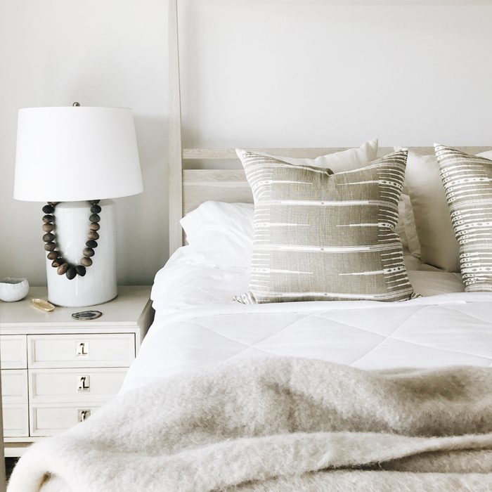 Bed with patterned pillows rests in a soft, neutral-toned bedroom beside a nightstand featuring a white lamp and decorative objects.