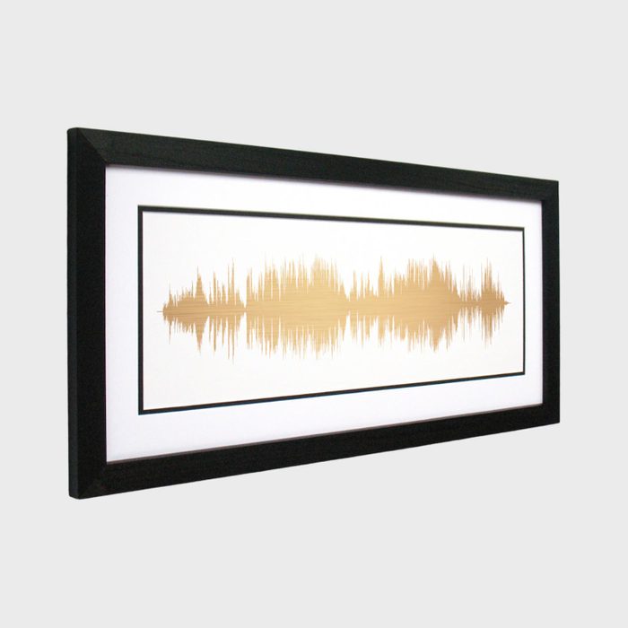 A framed artwork displays gold soundwaves on a white background, with a black border, set against a plain gray wall.