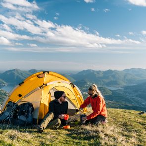 A yellow tent stands on a grassy hill; two people prepare drinks, overlooking distant mountains under a vibrant blue sky.